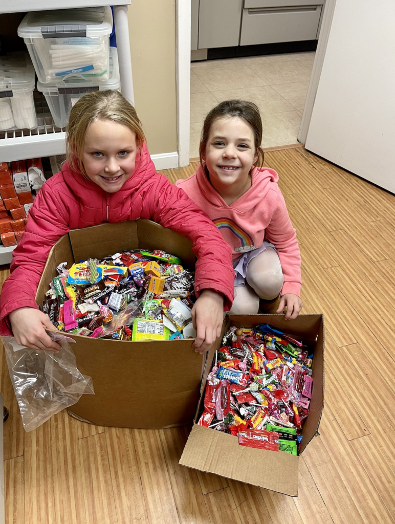 Two children smiling at the camera while lording over a cardboard box stuffed with candy.