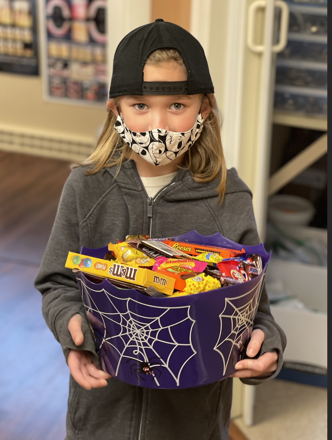 A child wearing a Jack Skellington mask holding a purple bucket of Halloween candy in both hands.